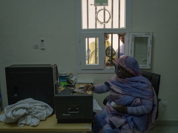 A woman uses a calculator at a desk as two people stand outside an open window.