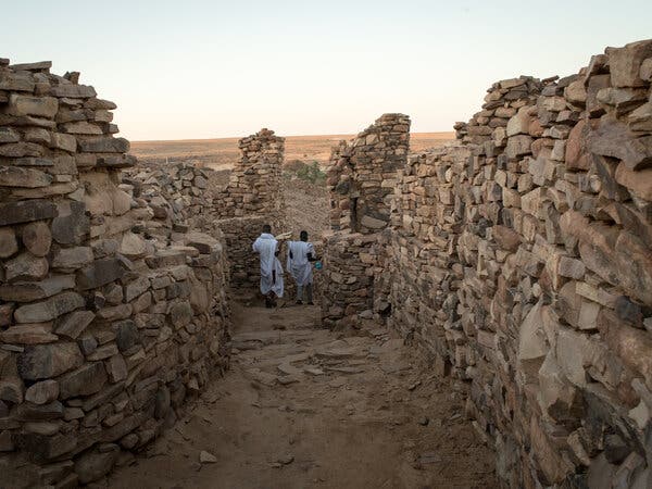 Two men carry loaves of bread in a walkway between tall stone walls.