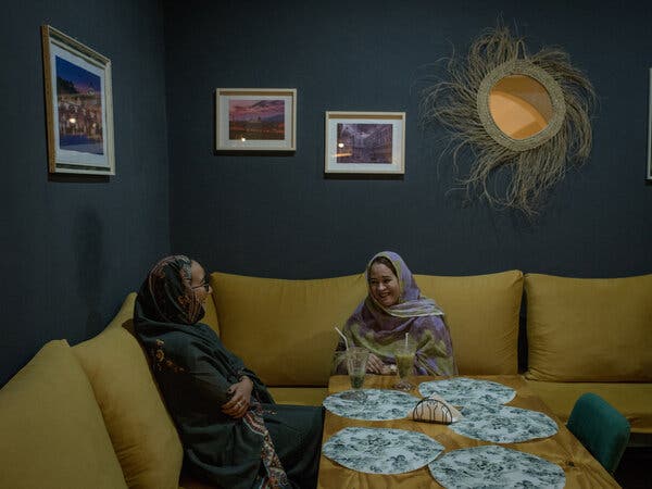 Two women sit on sofa-like seating at a table in a room adorned with pictures and a mirror.