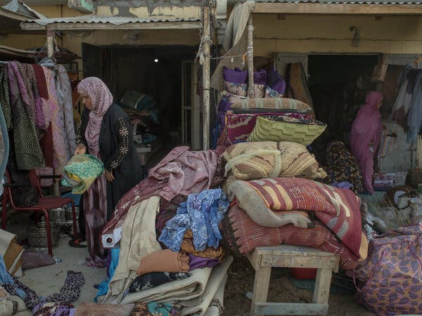 Two women stand amid stacks of fabrics, pillows and other things for sale.