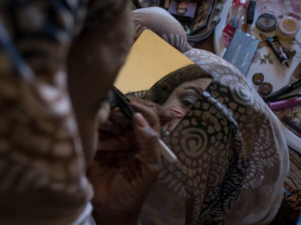 A woman applies makeup to her face while looking into a hand-held mirror.
