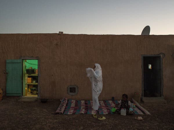 A person dressed in a white garment stands in a courtyard while s child sits on a rug behind her.