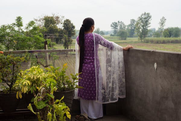 A woman in a purple outfit stands on a terrace, with her back to the camera. 