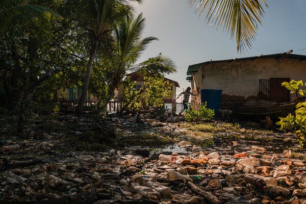 A person walking by an area filled with plastic trash. Several small buildings are standing on posts in the water.