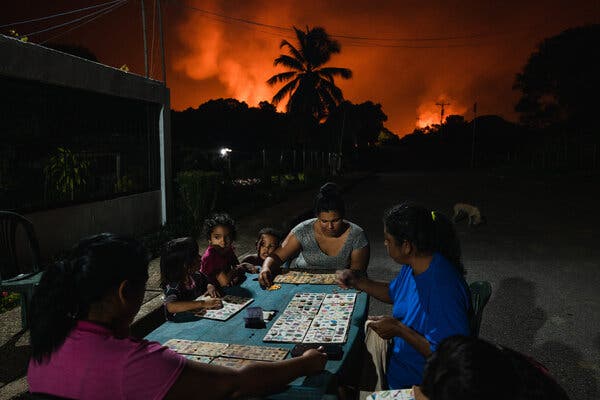 Adults playing board games at a table while children watch them. Behind them, gas flares are lighting up the night sky.