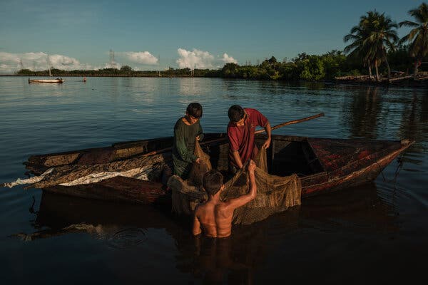 Three boys rummaging through a fishing net. Two are in a rickety boat, while the third is standing in the lake.