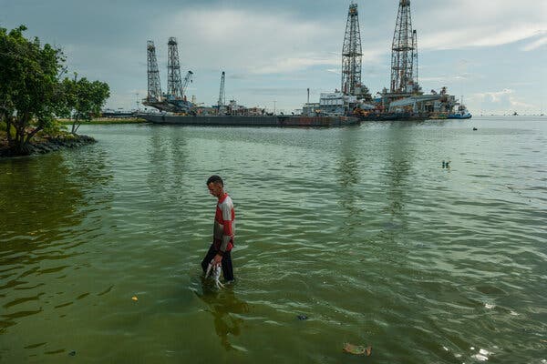 A man holding fish while standing in a green lake. Oil rigs are in the distance.