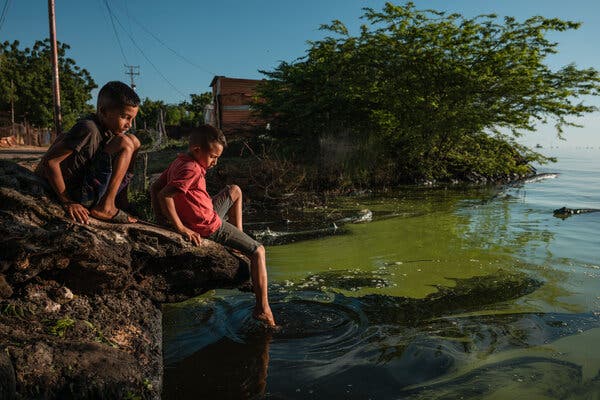 Two boys at the edge of a green lake on a sunny day. One boy is dipping his foot into the water while the other watches.