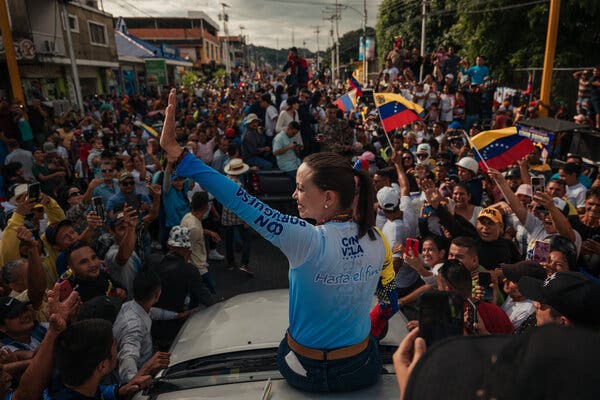 Ms. Machado sits on the roof of a car and waves to a crowd. 